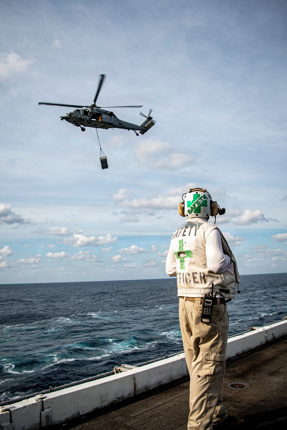A Safety Officer stands on the deck of a naval vessel observing a military helicopter conducting a vertical replenishment (VERTREP) operation over open ocean. The helicopter, likely an MH-60 Seahawk, is airborne with a cargo load suspended beneath it via a sling. The Safety Officer is wearing standard flight deck safety gear including a white vest, helmet with green cross markings, ear protection, and carries a radio.