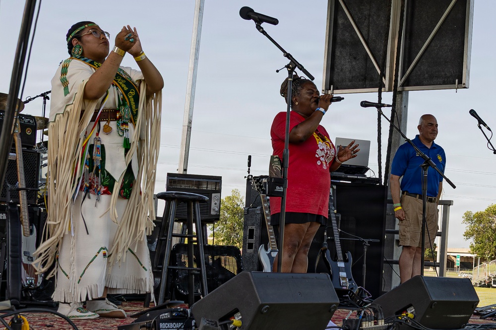 Three people are on an outdoor stage during what appears to be a cultural event or festival. A person on the left is wearing traditional Native American regalia with white fringe and beadwork, while a woman in a red shirt sings into a microphone at center stage. A man in a blue polo shirt stands to the right near another microphone.