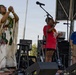 Three people are on an outdoor stage during what appears to be a cultural event or festival. A person on the left is wearing traditional Native American regalia with white fringe and beadwork, while a woman in a red shirt sings into a microphone at center stage. A man in a blue polo shirt stands to the right near another microphone.