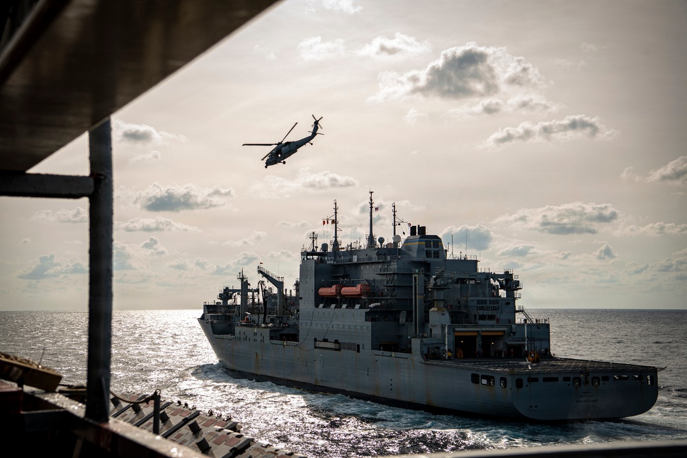 A military naval replenishment vessel is underway at sea, photographed from the deck of another ship. A military helicopter, consistent with an MH-60 Seahawk type, is flying above the replenishment ship. The scene appears to depict a replenishment-at-sea (RAS) or vertical replenishment (VERTREP) operation.