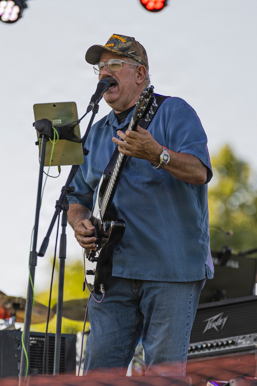 A middle-aged to older man is performing on an outdoor stage, playing an electric guitar and singing into a microphone. He is wearing a blue button-up shirt, jeans, a camouflage cap, and glasses, with a watch on his wrist. A tablet on a stand is positioned nearby, likely displaying lyrics or sheet music.