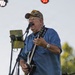 A middle-aged to older man is performing on an outdoor stage, playing an electric guitar and singing into a microphone. He is wearing a blue button-up shirt, jeans, a camouflage cap, and glasses, with a watch on his wrist. A tablet on a stand is positioned nearby, likely displaying lyrics or sheet music.