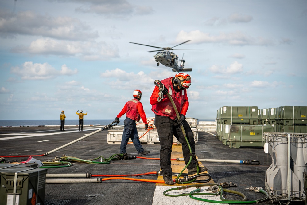 Military personnel in red uniforms and helmets are conducting flight deck operations aboard a naval vessel, managing fuel hoses and equipment while a helicopter hovers overhead. The scene depicts a coordinated replenishment or fueling operation on what appears to be an aircraft carrier or amphibious assault ship flight deck. Signal personnel in yellow vests are visible in the background directing the helicopter.