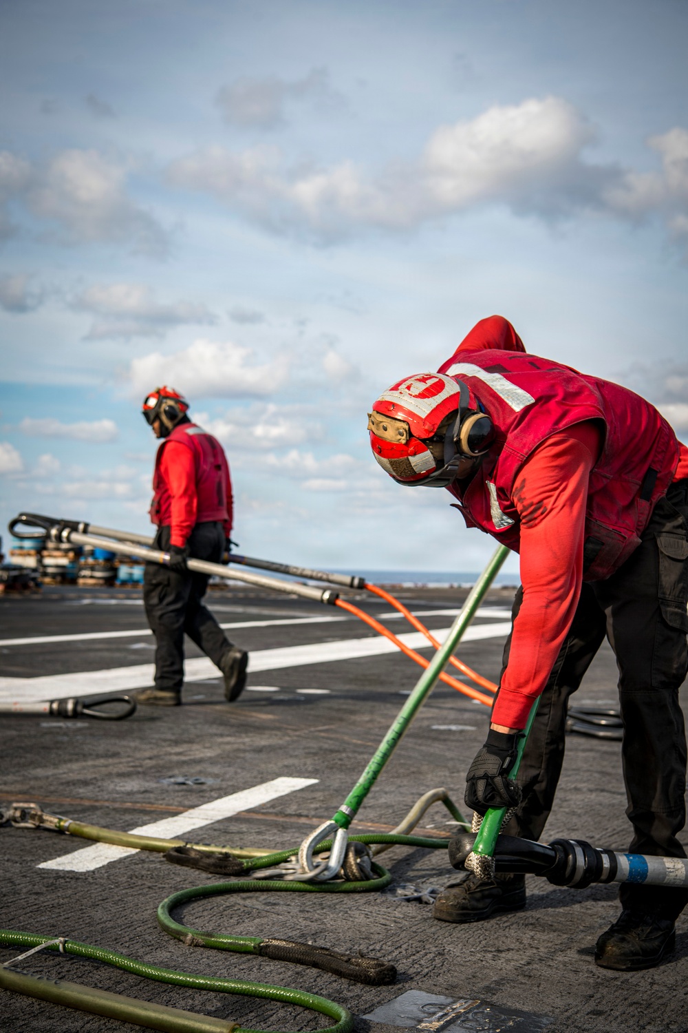 Two personnel dressed in red flight deck jerseys and helmets are working on what appears to be an aircraft carrier flight deck. The foreground worker is bent over handling green fueling hoses and equipment, while the background worker walks carrying a long metal pole or rod. The scene depicts routine flight deck operations, likely involving aircraft refueling or deck maintenance.