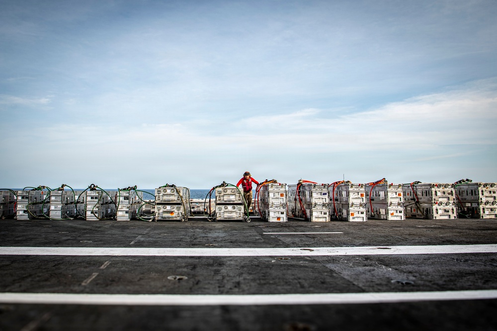 A person in a red jacket stands among a long row of military supply containers or equipment crates arranged on what appears to be the flight deck of a naval vessel. The containers are secured with straps and hoses, suggesting they are ready for transport or have recently been delivered via replenishment at sea. The ocean horizon is visible in the background, confirming the maritime setting.