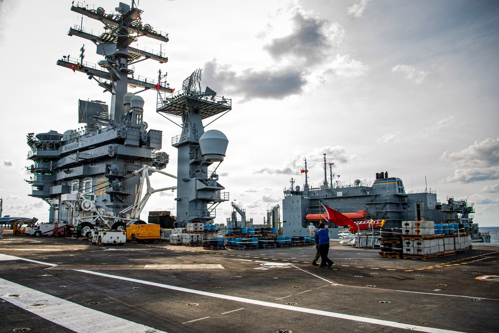 The image shows the flight deck of a large US Navy aircraft carrier during an underway replenishment (UNREP) operation with a supply vessel visible alongside to starboard. Numerous pallets of supplies and equipment are staged on the flight deck, and the carrier's island superstructure with radar arrays and communication equipment is prominently visible on the left. Two personnel in blue uniforms are walking across the deck amid the ongoing logistics operation.