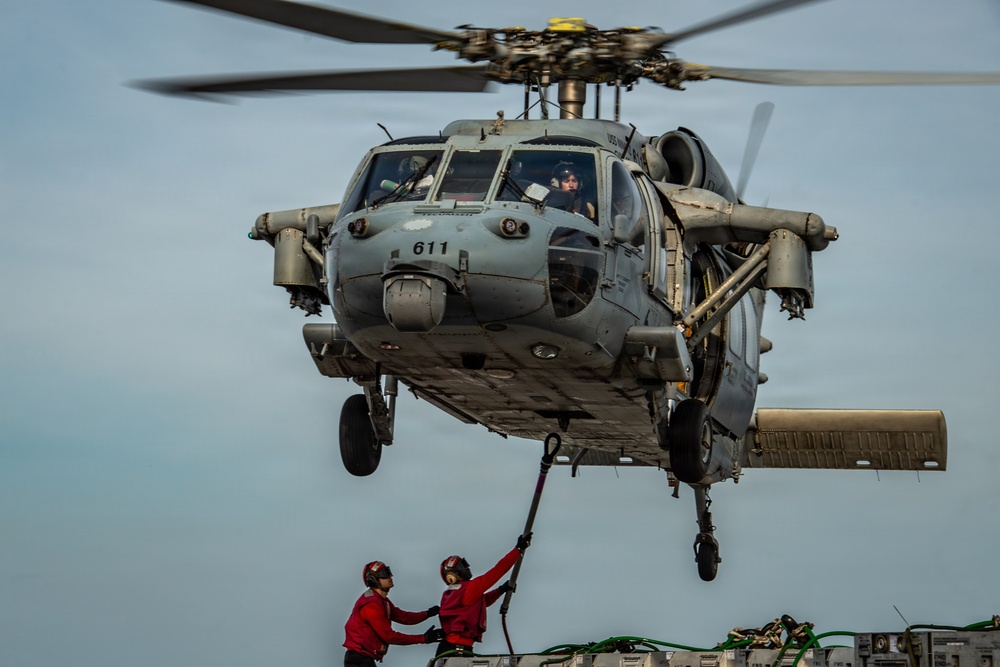 A military helicopter numbered '611' hovers low over what appears to be a naval vessel deck. Two personnel dressed in red flight deck gear are reaching up toward a cable or hose connected to the aircraft, likely performing a helicopter in-flight refueling (HIFR) or cargo transfer operation. The image is taken from a close, low-angle perspective that emphasizes the scale and proximity of the hovering aircraft.