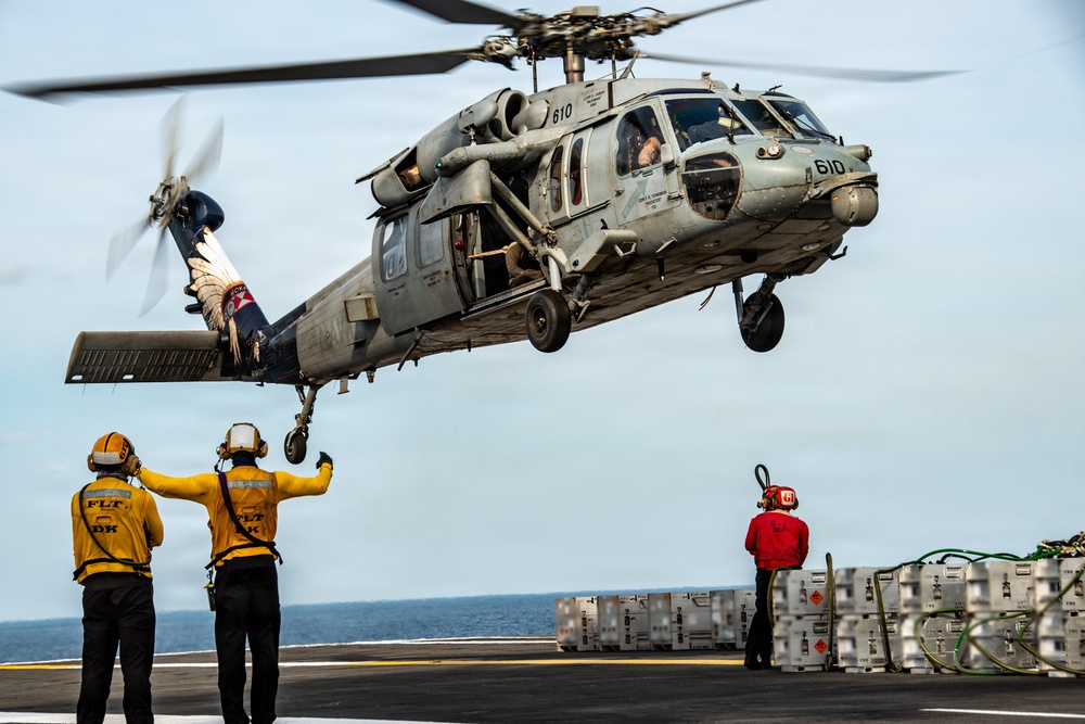 A U.S. Navy MH-60S Seahawk helicopter (bureau number 610) hovers low over the flight deck of a naval vessel during what appears to be a vertical replenishment or deck operations exercise. Three flight deck crew members in yellow and red jerseys are visible, with two signaling the helicopter and one standing near cargo containers. The scene takes place at sea under overcast skies.