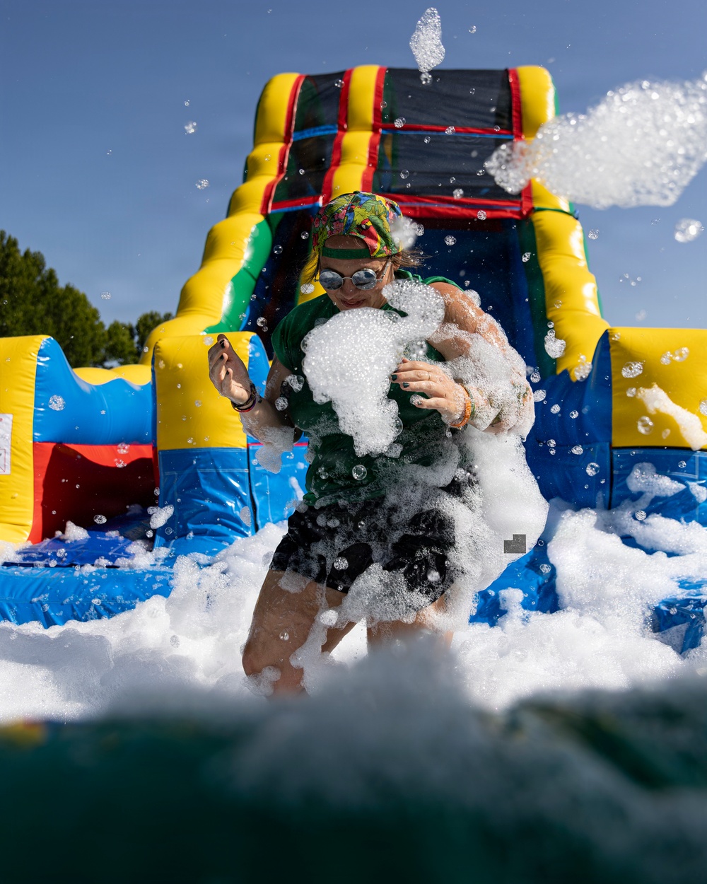 A person is participating in a foam party or foam run event, standing in front of a large colorful inflatable bounce house/water slide. They are covered in white soapy foam with bubbles flying through the air around them. The photo is taken from a low angle on a clear sunny day, capturing the dynamic and playful atmosphere of the event.