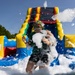 A person is participating in a foam party or foam run event, standing in front of a large colorful inflatable bounce house/water slide. They are covered in white soapy foam with bubbles flying through the air around them. The photo is taken from a low angle on a clear sunny day, capturing the dynamic and playful atmosphere of the event.
