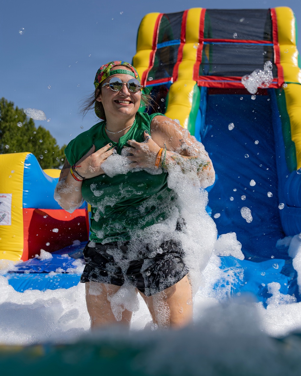 A smiling woman is playing in a foam/bubble pit at an outdoor event on a sunny day. She is covered in white foam suds and appears to be having an enthusiastic, joyful time. Behind her is a colorful inflatable bounce house and water slide structure.