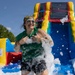 A smiling woman is playing in a foam/bubble pit at an outdoor event on a sunny day. She is covered in white foam suds and appears to be having an enthusiastic, joyful time. Behind her is a colorful inflatable bounce house and water slide structure.