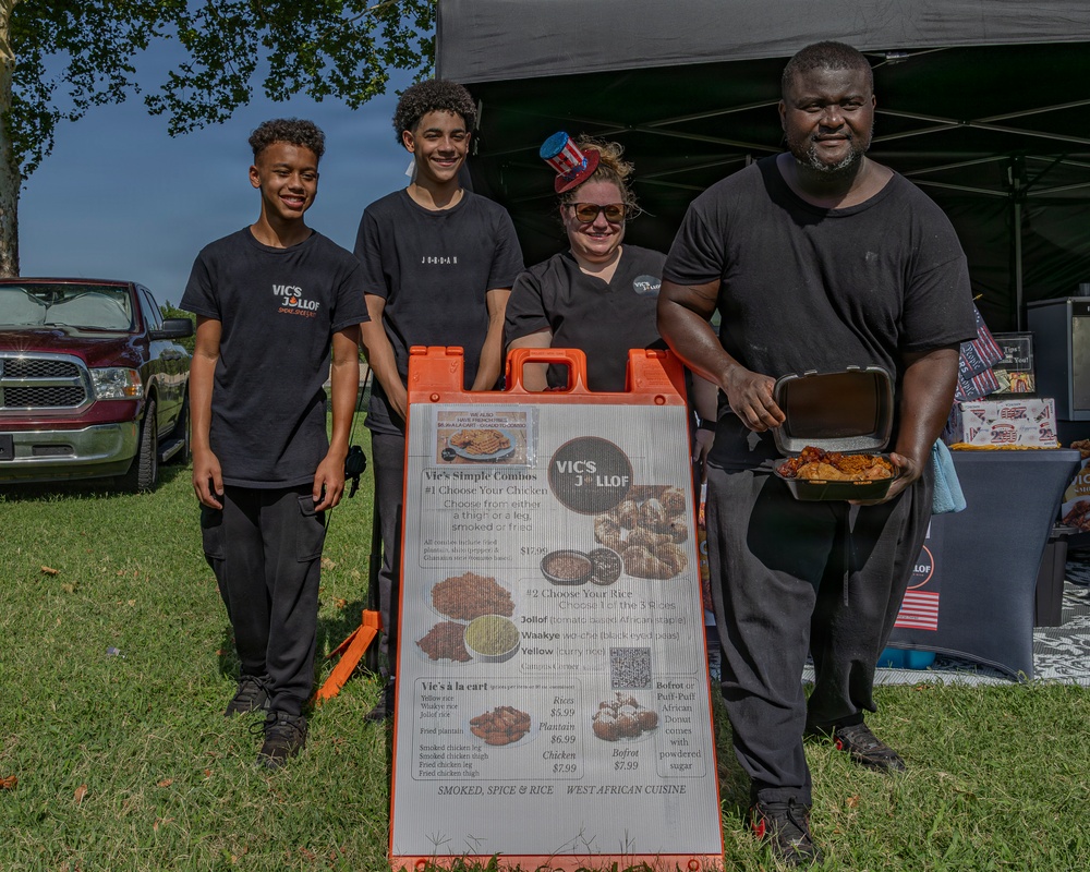 Four people pose together at an outdoor food vendor booth for 'Vic's Jollof,' a West African cuisine business. They are standing in front of a sandwich board menu sign displaying combo meals and à la carte options. The setting appears to be an outdoor festival or community event on a sunny day.