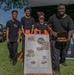 Four people pose together at an outdoor food vendor booth for 'Vic's Jollof,' a West African cuisine business. They are standing in front of a sandwich board menu sign displaying combo meals and à la carte options. The setting appears to be an outdoor festival or community event on a sunny day.
