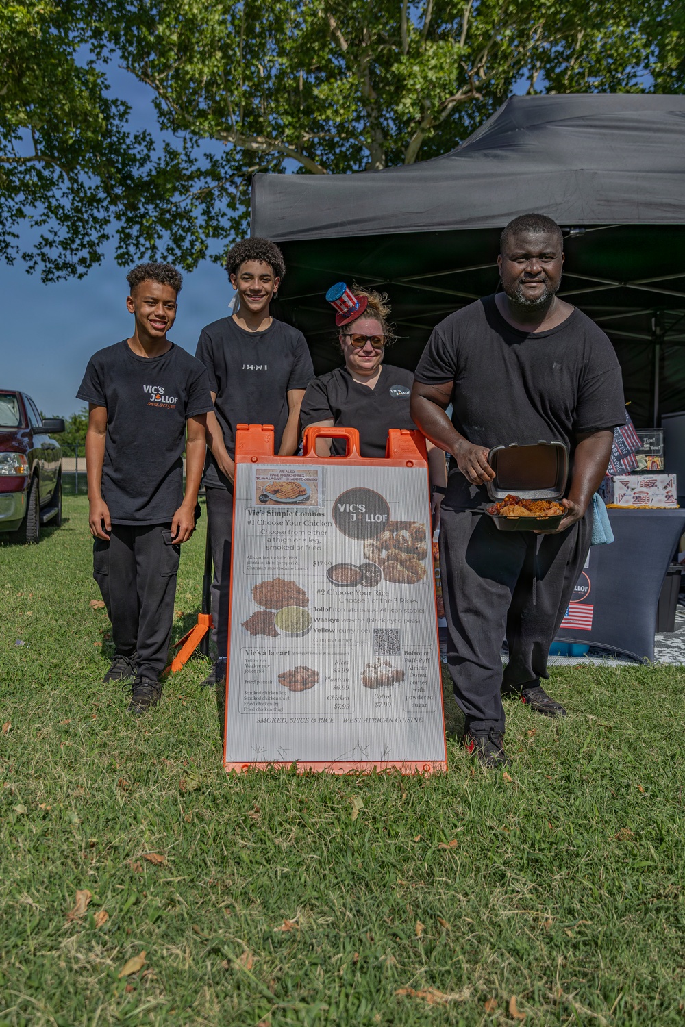 Four people pose together at an outdoor food vendor booth for 'Vic's Jollof,' a West African cuisine business. They are standing in front of an A-frame menu board displaying combo options and pricing, set up under a black canopy tent on a grassy area. The group appears to be a family or team operating the food stall at what looks like an outdoor festival or market event.