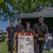 Four people pose together at an outdoor food vendor booth for 'Vic's Jollof,' a West African cuisine business. They are standing in front of an A-frame menu board displaying combo options and pricing, set up under a black canopy tent on a grassy area. The group appears to be a family or team operating the food stall at what looks like an outdoor festival or market event.