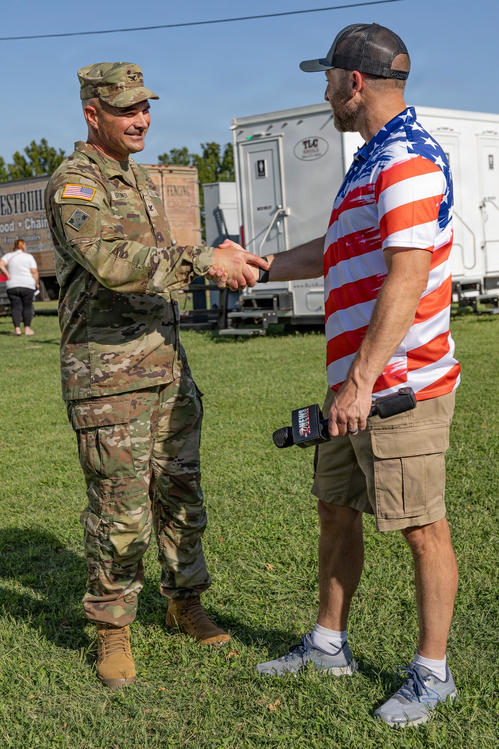 A U.S. Army soldier in OCP (Operational Camouflage Pattern) uniform shakes hands with a civilian wearing an American flag-themed shirt at an outdoor event. The civilian is holding a microphone branded with what appears to be a news station logo, suggesting a media interview or public event. The setting appears to be a community or public gathering on a grassy field.