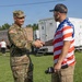A U.S. Army soldier in OCP (Operational Camouflage Pattern) uniform shakes hands with a civilian wearing an American flag-themed shirt at an outdoor event. The civilian is holding a microphone branded with what appears to be a news station logo, suggesting a media interview or public event. The setting appears to be a community or public gathering on a grassy field.