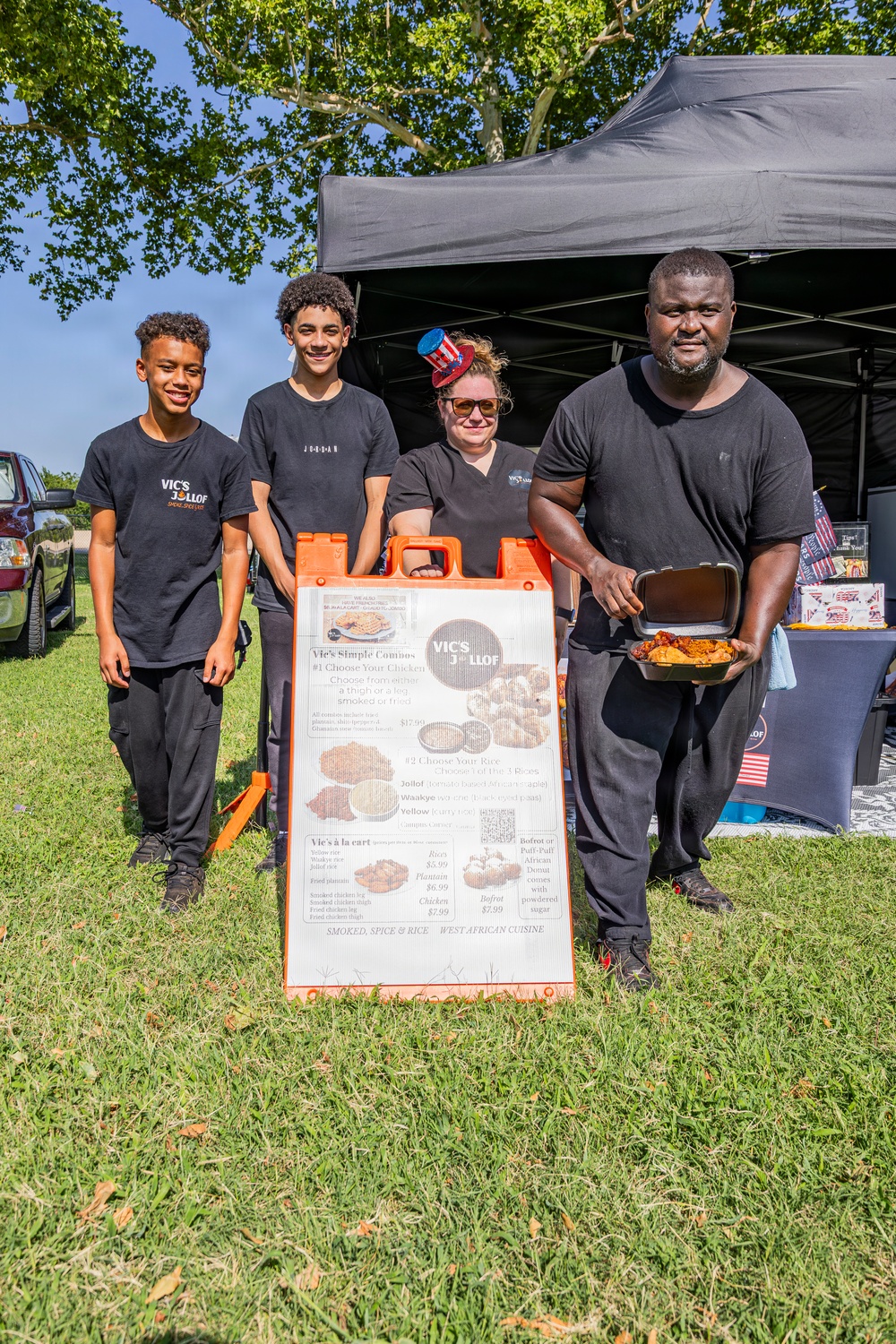 Four people pose together at an outdoor food vendor booth called 'Vic's Jollof,' a West African cuisine stand. The group includes two teenage boys, a woman wearing a patriotic mini top hat and sunglasses, and a man holding an open food container with what appears to be chicken and rice. They are set up under a black canopy tent at what appears to be an outdoor festival or market.