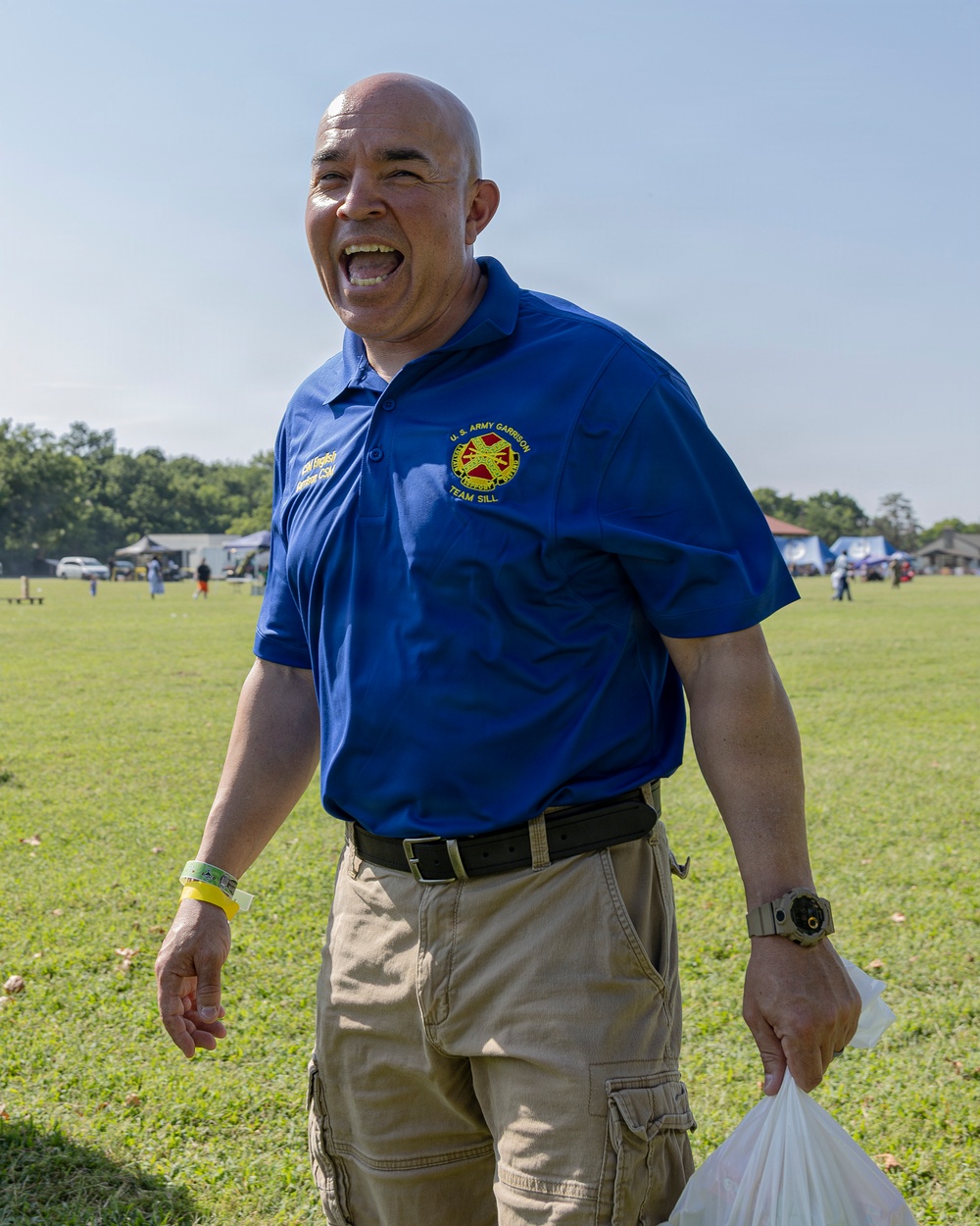 A bald man is laughing enthusiastically outdoors at what appears to be a community or military event. He is wearing a blue polo shirt with a 'U.S. Army Garrison Team Sill' emblem and a name tag, along with khaki cargo shorts. He is holding a plastic bag in his right hand and wearing a yellow wristband and a watch.