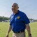 A bald man is laughing enthusiastically outdoors at what appears to be a community or military event. He is wearing a blue polo shirt with a 'U.S. Army Garrison Team Sill' emblem and a name tag, along with khaki cargo shorts. He is holding a plastic bag in his right hand and wearing a yellow wristband and a watch.