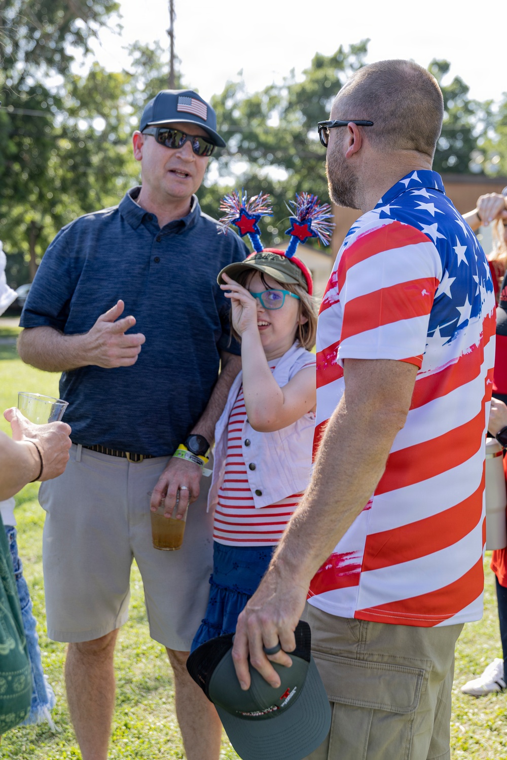 Three individuals are gathered outdoors at what appears to be a patriotic celebration, likely a Fourth of July event. A man in a navy polo with an American flag cap, a child wearing festive patriotic accessories, and a man in an American flag-patterned shirt are engaged in conversation. The setting is a grassy outdoor area with trees visible in the background.