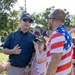 Three individuals are gathered outdoors at what appears to be a patriotic celebration, likely a Fourth of July event. A man in a navy polo with an American flag cap, a child wearing festive patriotic accessories, and a man in an American flag-patterned shirt are engaged in conversation. The setting is a grassy outdoor area with trees visible in the background.