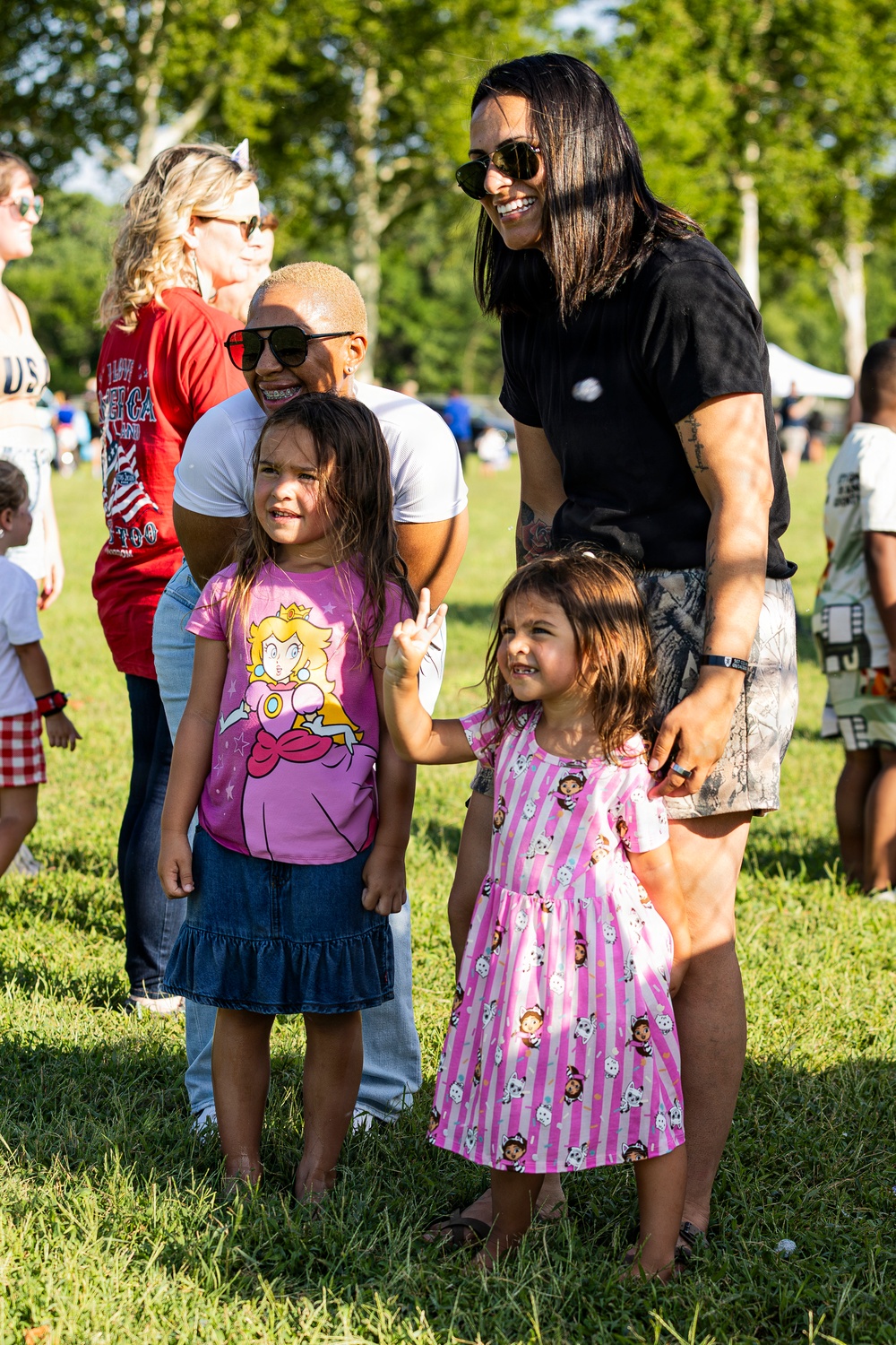 A group of adults and two young girls are gathered outdoors on a grassy field, appearing to pose for a photo at what looks like a community or family event. The two children are dressed in colorful character-themed clothing, with one wearing a Princess Peach shirt and denim skirt, and the other in a pink striped dress. The adults are smiling and appear to be enjoying the outdoor gathering.