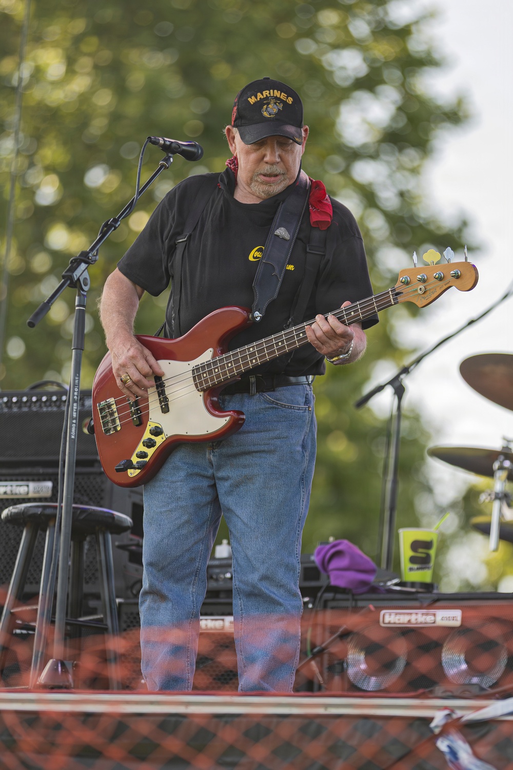 An older male musician is performing outdoors on a stage, playing a red electric bass guitar. He is wearing a black 'Marines' cap, a black t-shirt, blue jeans, and a red bandana around his neck. The setting appears to be an outdoor concert with trees visible in the background and music equipment surrounding him.