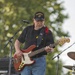 An older male musician is performing outdoors on a stage, playing a red electric bass guitar. He is wearing a black 'Marines' cap, a black t-shirt, blue jeans, and a red bandana around his neck. The setting appears to be an outdoor concert with trees visible in the background and music equipment surrounding him.