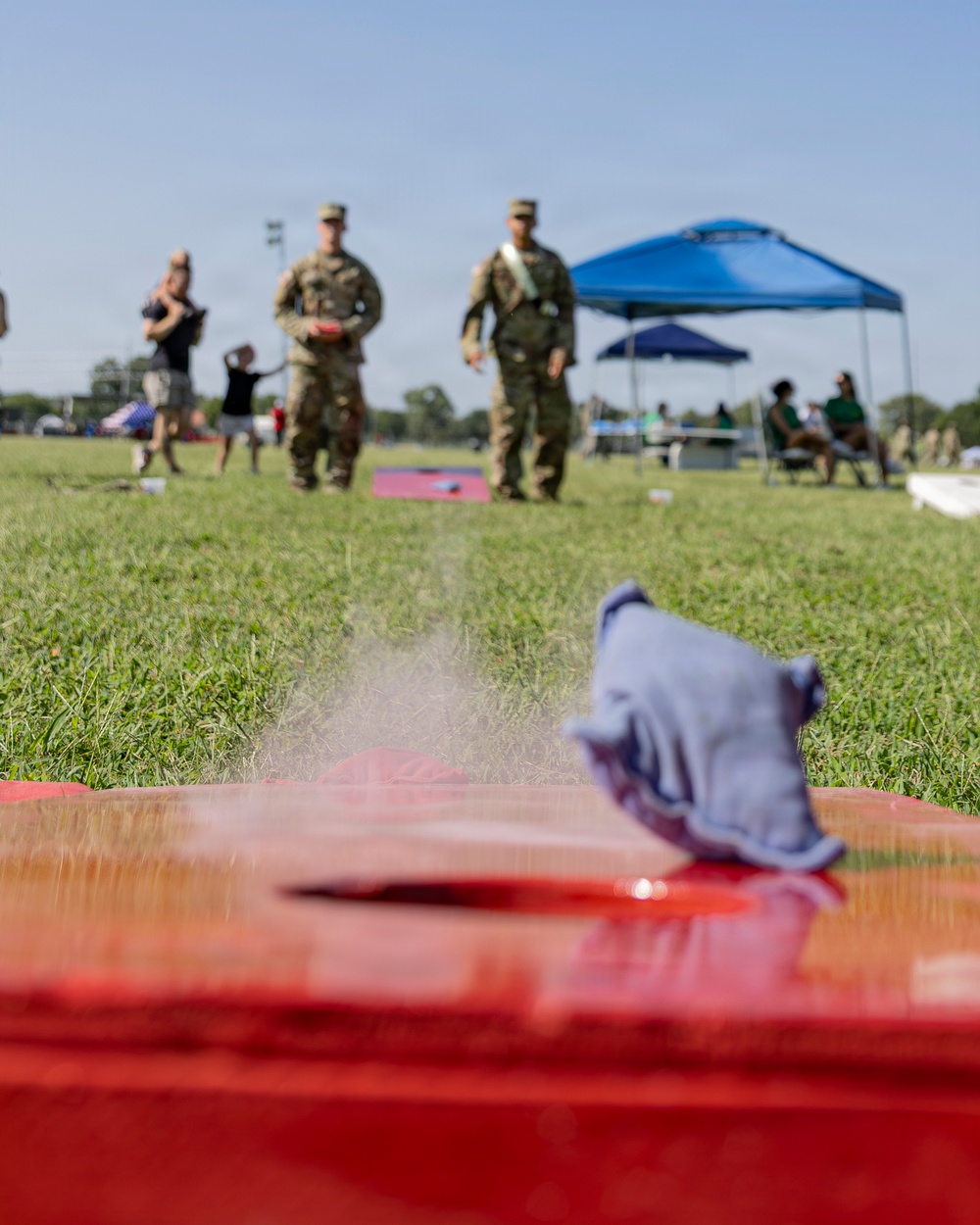 A ground-level perspective shot of a cornhole game being played at an outdoor military gathering or event. Two soldiers in camouflage uniforms stand at the far end of the board, with a bean bag visible mid-slide on the red wooden board in the foreground. The event appears to be a casual recreational gathering with families, tents, and other attendees visible in the background.