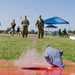 A ground-level perspective shot of a cornhole game being played at an outdoor military gathering or event. Two soldiers in camouflage uniforms stand at the far end of the board, with a bean bag visible mid-slide on the red wooden board in the foreground. The event appears to be a casual recreational gathering with families, tents, and other attendees visible in the background.