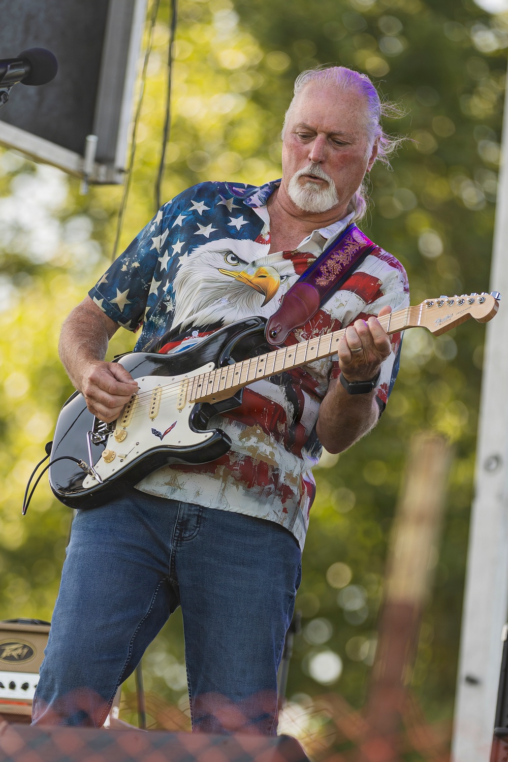 An older white-haired man with a goatee is performing on an outdoor stage, playing an electric guitar with focused concentration. He is wearing a patriotic American flag shirt featuring a bald eagle graphic and blue jeans. The setting appears to be an outdoor music festival or concert during daylight hours.