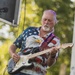 An older white-haired man with a goatee is performing on an outdoor stage, playing an electric guitar with focused concentration. He is wearing a patriotic American flag shirt featuring a bald eagle graphic and blue jeans. The setting appears to be an outdoor music festival or concert during daylight hours.