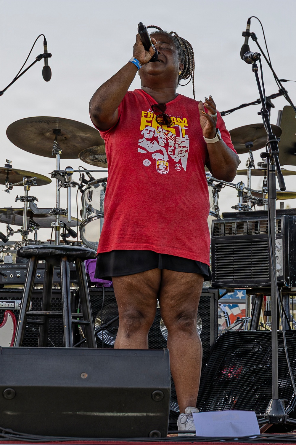 A woman is performing on an outdoor stage, singing into a handheld microphone with her eyes closed and head tilted back. She is wearing a red 'Freedom Fest' t-shirt featuring Mount Rushmore imagery and black shorts. A full drum kit, cymbals, monitor speakers, and other stage equipment are visible behind her.
