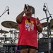 A woman is performing on an outdoor stage, singing into a handheld microphone with her eyes closed and head tilted back. She is wearing a red 'Freedom Fest' t-shirt featuring Mount Rushmore imagery and black shorts. A full drum kit, cymbals, monitor speakers, and other stage equipment are visible behind her.
