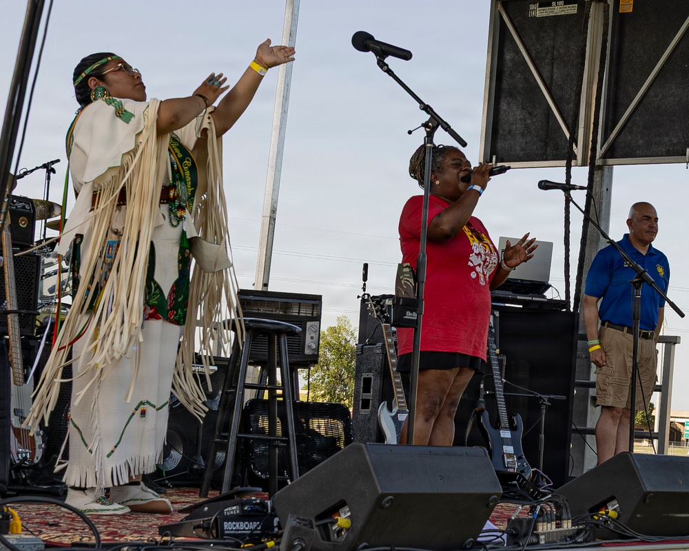 An outdoor stage performance featuring three individuals, with two actively performing. A person in traditional Native American regalia with white fringe garments raises their arm expressively, while a woman in a red shirt sings passionately into a microphone. A third person in a blue polo shirt stands to the right near another microphone stand.