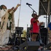 An outdoor stage performance featuring three individuals, with two actively performing. A person in traditional Native American regalia with white fringe garments raises their arm expressively, while a woman in a red shirt sings passionately into a microphone. A third person in a blue polo shirt stands to the right near another microphone stand.
