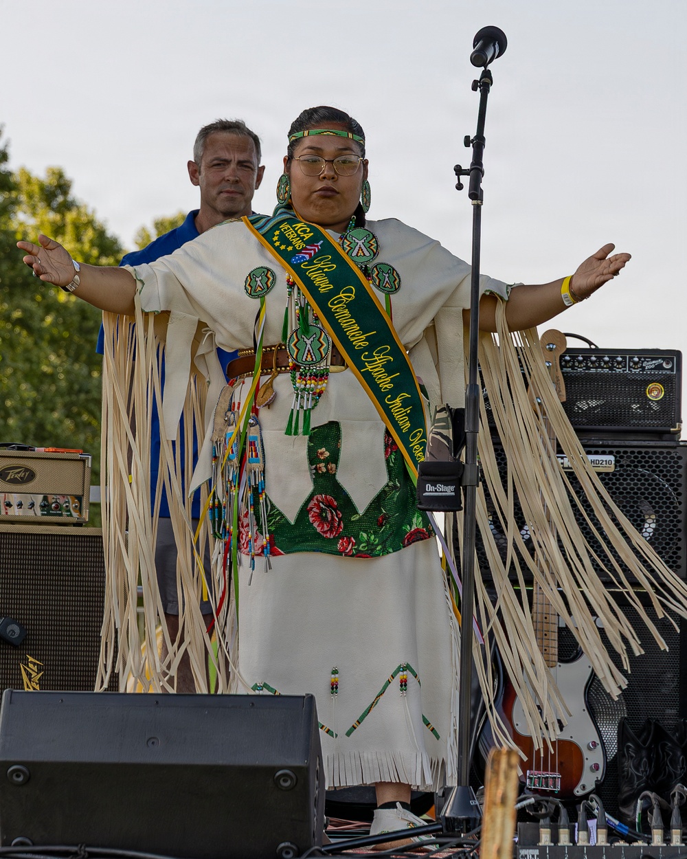 A young woman wearing traditional Native American regalia stands on an outdoor stage with arms outstretched. She is wearing a green and gold sash that reads 'KCA Veterans Kiowa Comanche Apache Indian Queen' along with a fringed white dress, beaded jewelry, and a beaded headband. Behind her stands a man in a blue polo shirt, and the stage is surrounded by musical equipment including amplifiers and a guitar.