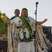 A young woman wearing traditional Native American regalia stands on an outdoor stage with arms outstretched. She is wearing a green and gold sash that reads 'KCA Veterans Kiowa Comanche Apache Indian Queen' along with a fringed white dress, beaded jewelry, and a beaded headband. Behind her stands a man in a blue polo shirt, and the stage is surrounded by musical equipment including amplifiers and a guitar.