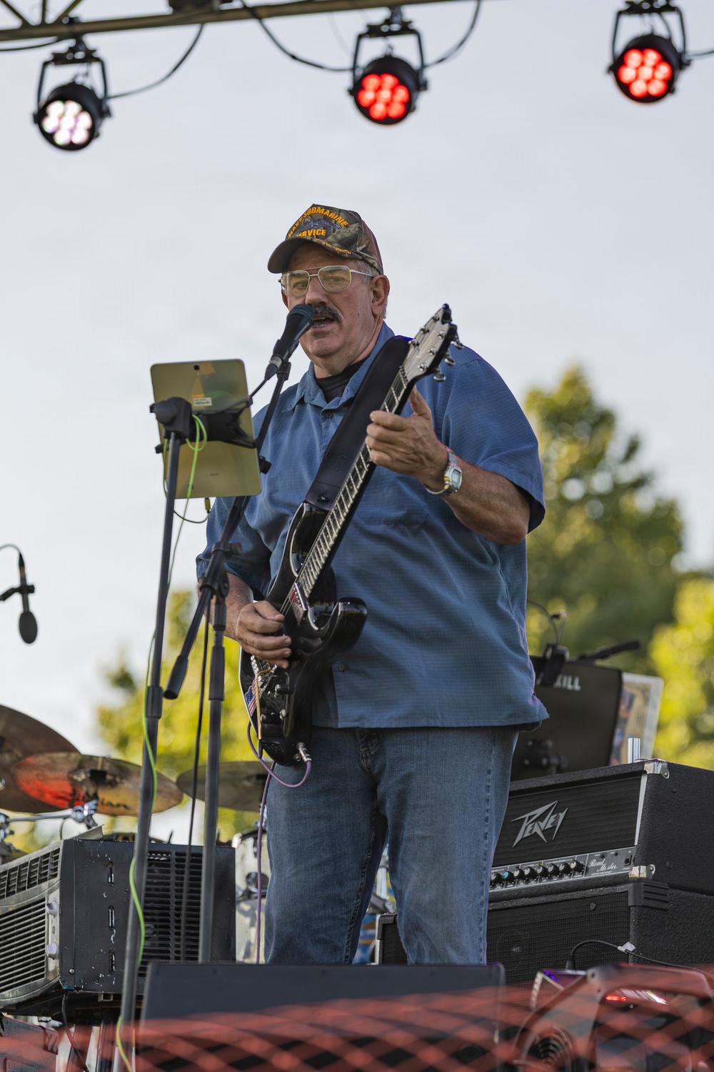 A middle-aged male guitarist performs on an outdoor stage during what appears to be a daytime or early evening event. He is singing into a microphone while playing a black electric guitar, wearing a military-themed cap, glasses, and a blue short-sleeve shirt. Stage lighting rigs are visible overhead along with a Peavey amplifier in the background.