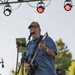 A middle-aged male guitarist performs on an outdoor stage during what appears to be a daytime or early evening event. He is singing into a microphone while playing a black electric guitar, wearing a military-themed cap, glasses, and a blue short-sleeve shirt. Stage lighting rigs are visible overhead along with a Peavey amplifier in the background.