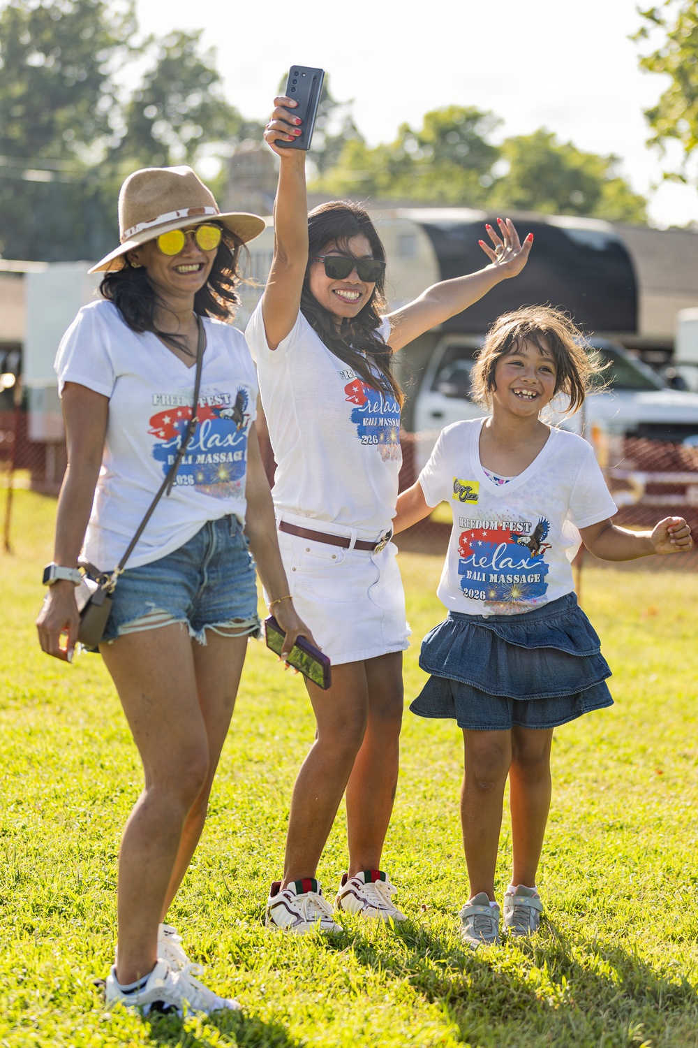 Three individuals — two adult women and a young girl — are posing and smiling outdoors on a grassy field at what appears to be a festival or outdoor event. All three are wearing matching white 'Freedom Fest / Relax Bali Massage 2026' promotional t-shirts. The group appears cheerful and celebratory, with one woman raising a smartphone in the air.