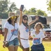 Three individuals — two adult women and a young girl — are posing and smiling outdoors on a grassy field at what appears to be a festival or outdoor event. All three are wearing matching white 'Freedom Fest / Relax Bali Massage 2026' promotional t-shirts. The group appears cheerful and celebratory, with one woman raising a smartphone in the air.