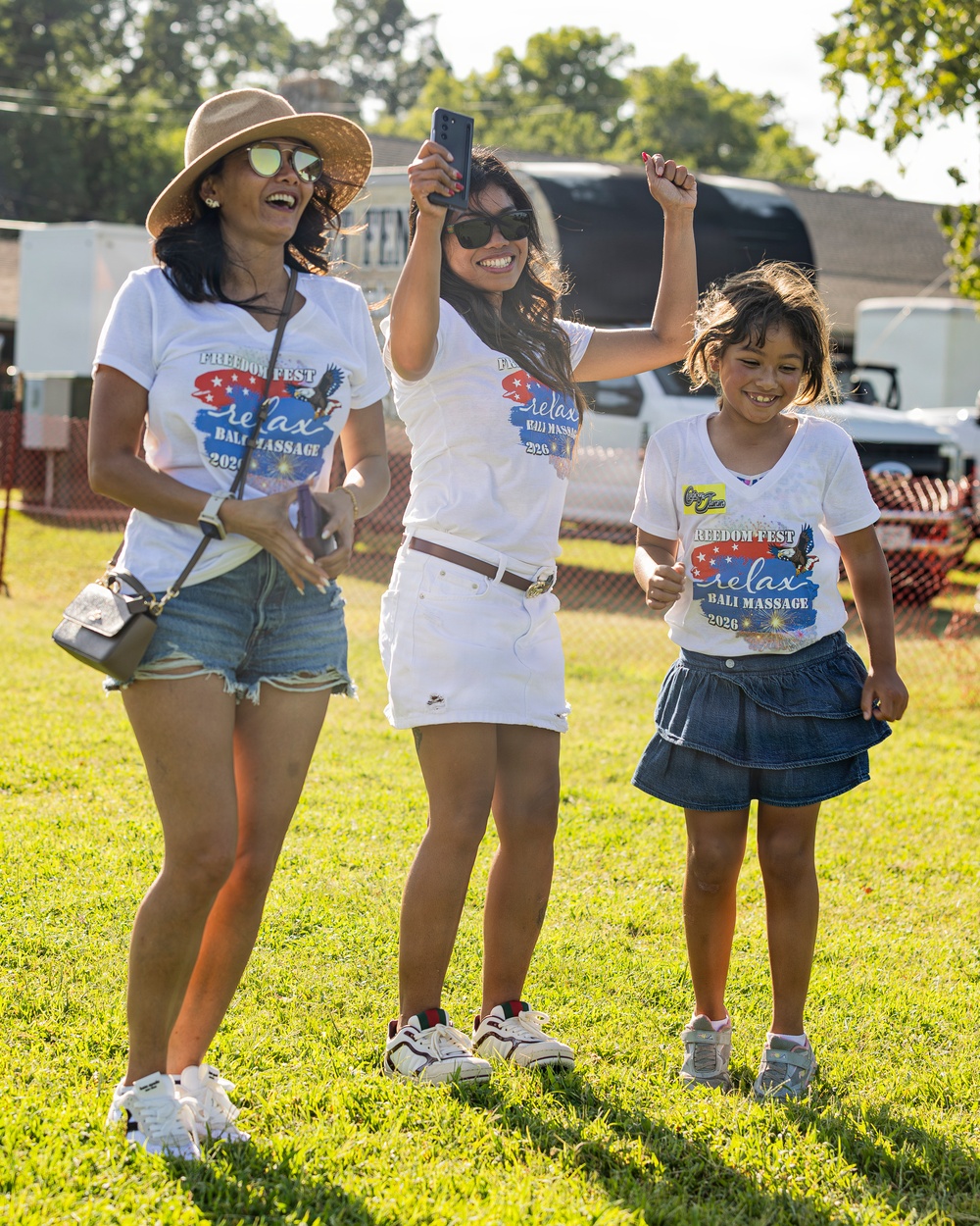 Three individuals — two adult women and a young girl — stand on a grassy lawn at an outdoor event. All three are wearing matching white 'Freedom Fest / Relax Bali Massage 2026' t-shirts, suggesting they are affiliated with or sponsored by the same organization. The group appears cheerful and celebratory, with one woman holding a smartphone raised in the air.