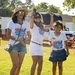 Three individuals — two adult women and a young girl — stand on a grassy lawn at an outdoor event. All three are wearing matching white 'Freedom Fest / Relax Bali Massage 2026' t-shirts, suggesting they are affiliated with or sponsored by the same organization. The group appears cheerful and celebratory, with one woman holding a smartphone raised in the air.