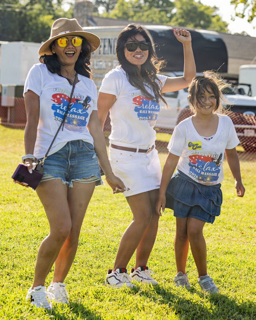 Three individuals — two adult women and a young girl — are posing and smiling on a grassy field at an outdoor event. All three are wearing matching white 'Freedom Fest / Relax Bali Massage 2026' event t-shirts. The mood is festive and celebratory, with one woman raising her fist in the air.