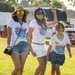 Three individuals — two adult women and a young girl — are posing and smiling on a grassy field at an outdoor event. All three are wearing matching white 'Freedom Fest / Relax Bali Massage 2026' event t-shirts. The mood is festive and celebratory, with one woman raising her fist in the air.