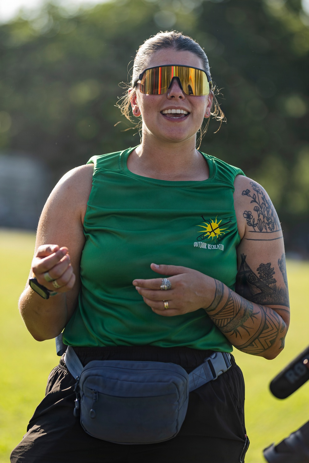 A smiling woman stands outdoors wearing a green 'Outdoor Recreation' sleeveless shirt, large mirrored visor-style sunglasses, and a gray fanny pack. She has extensive tattoos on her right arm featuring floral and geometric designs. The background shows a grassy field with blurred trees, suggesting an outdoor recreational event.