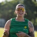 A smiling woman stands outdoors wearing a green 'Outdoor Recreation' sleeveless shirt, large mirrored visor-style sunglasses, and a gray fanny pack. She has extensive tattoos on her right arm featuring floral and geometric designs. The background shows a grassy field with blurred trees, suggesting an outdoor recreational event.